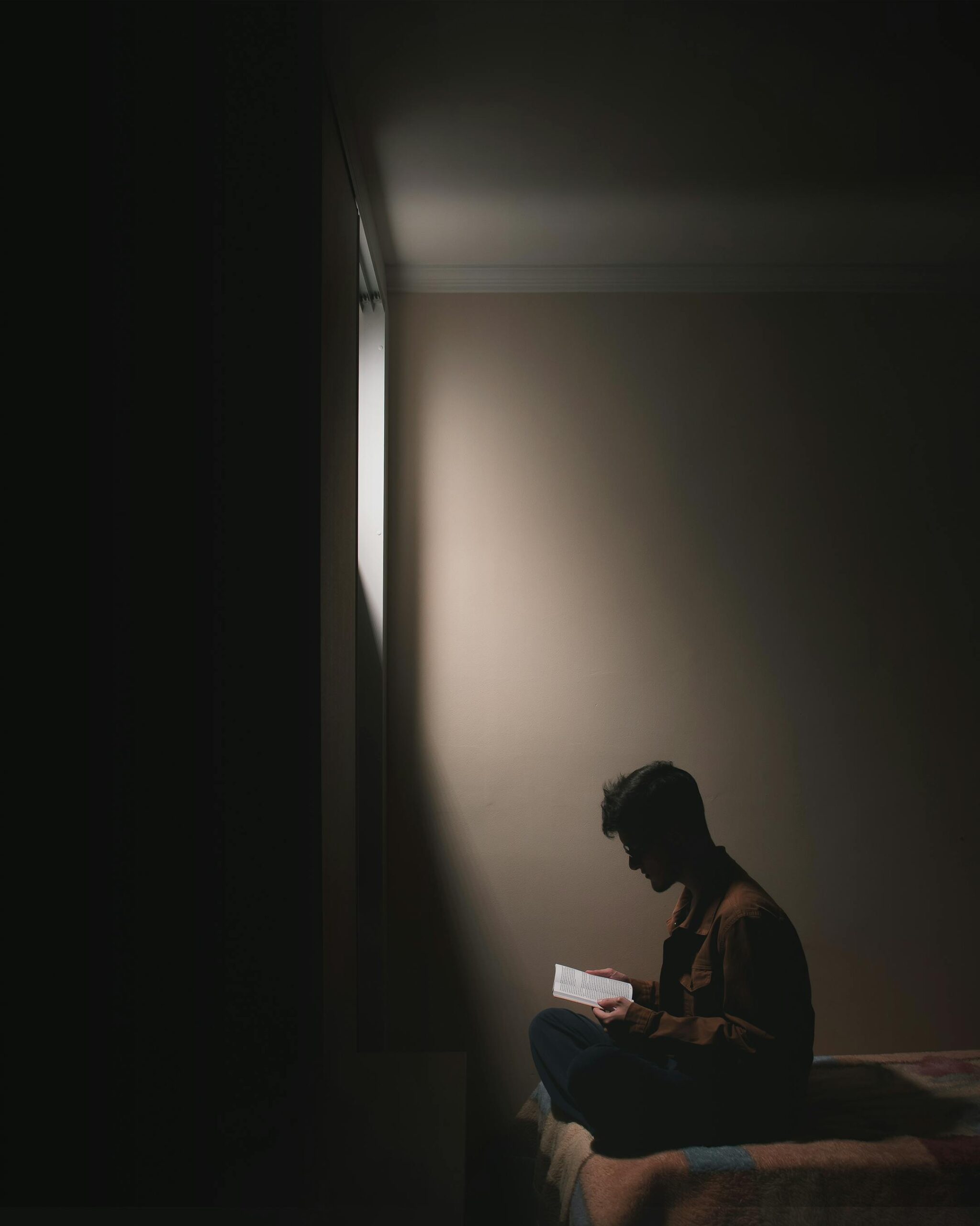 Silhouette of a man sitting on a bed in a dark room, reading a book by window light.